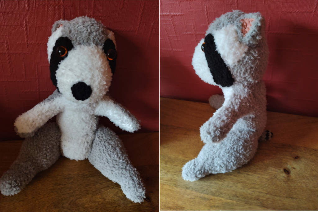 A fluffy crochet raccoon/bear sitting on a wooden surface in front of a red wall. The raccoon is shown from the front view on the left and the side view on the right It has grey body fur and a white front and face with black stripes over its orange eyes