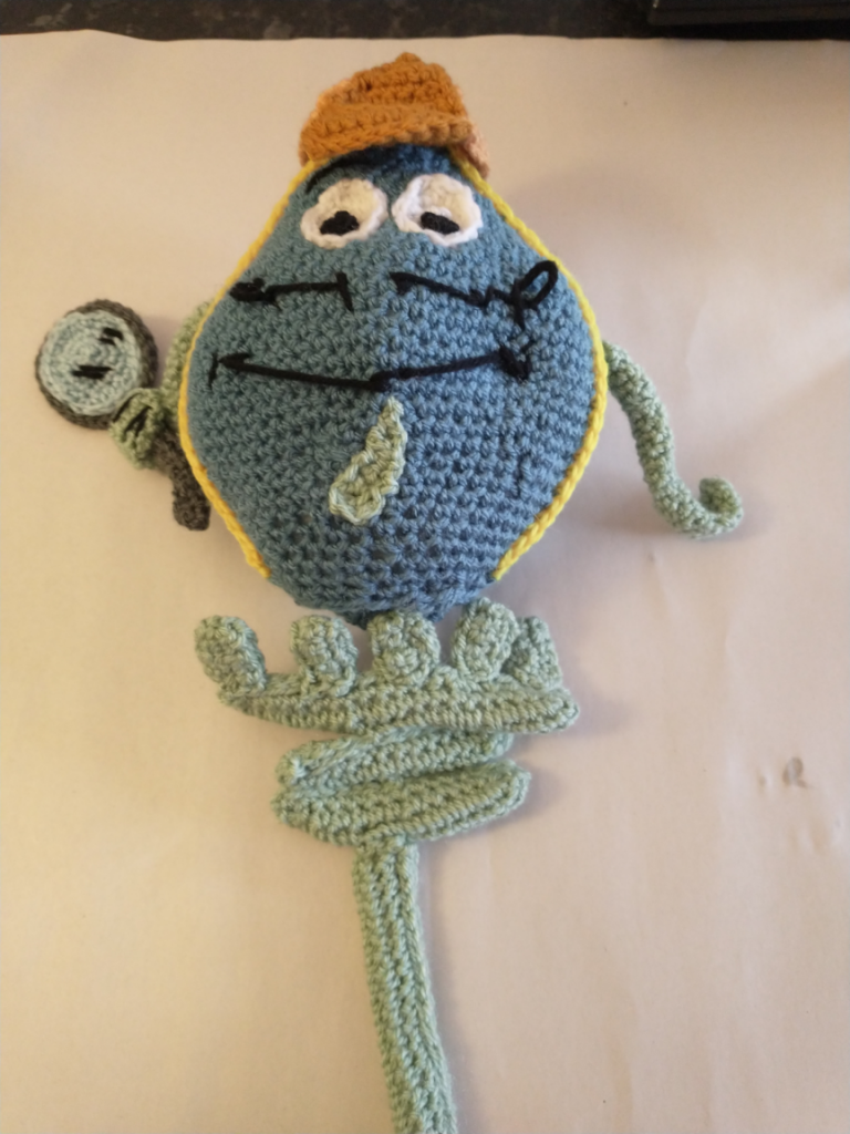 A crochet toy that is a raindrop shaped detective with a blue face and curly black moustache. he is wearing a tan deerstalker hat and is carrying a magnifying glass.