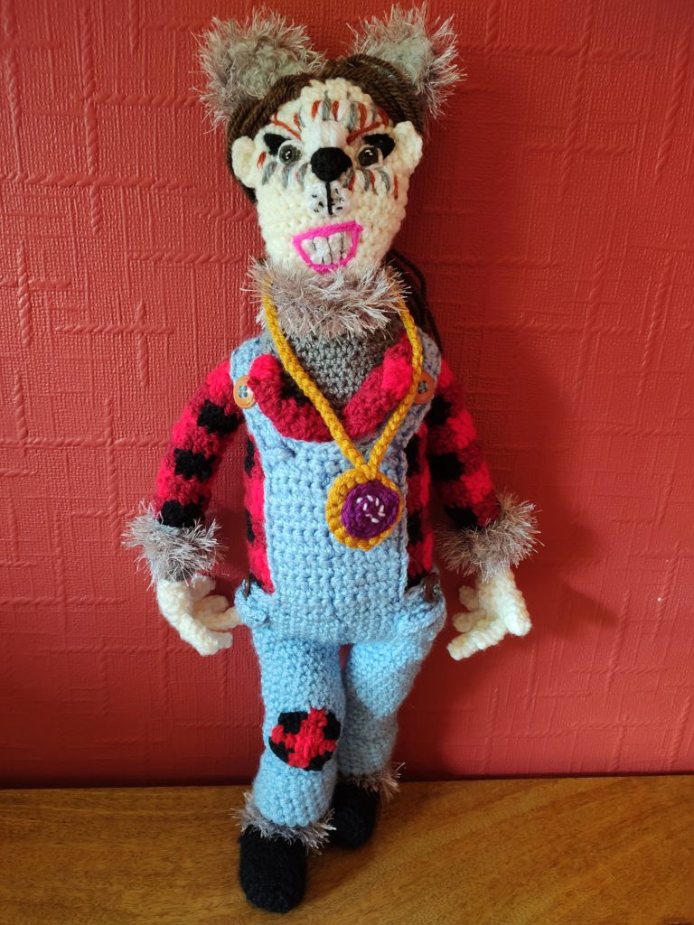 A photo of crochet doll of a little girl in her scary wolf dancing costume, which is a pair of blue dungarees over a red plaid shirt and grey undershirt with grey fur around the neck and wrists. She is wearing black dancing shoes and a pair of furry grey wolf ears. He long brown hair is pulled back and her face is painted to look like a wolf. She is wearing a medal around her neck and is baring her teeth like a wolf. She has a plaid patch on the right knee