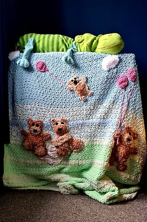 A baby blanket with teddy bears having a picnic on it. The bottom of the blanket is green striped with cream and the top had blue striped with cream. There are two large teddies in the middle on a picnic blanket and one large teddy on the right holding two pink balloons. A small teddy is in the sky being carried away by another pink balloon. The blanket is draped over a chest of drawers with a green hippo toy on top.