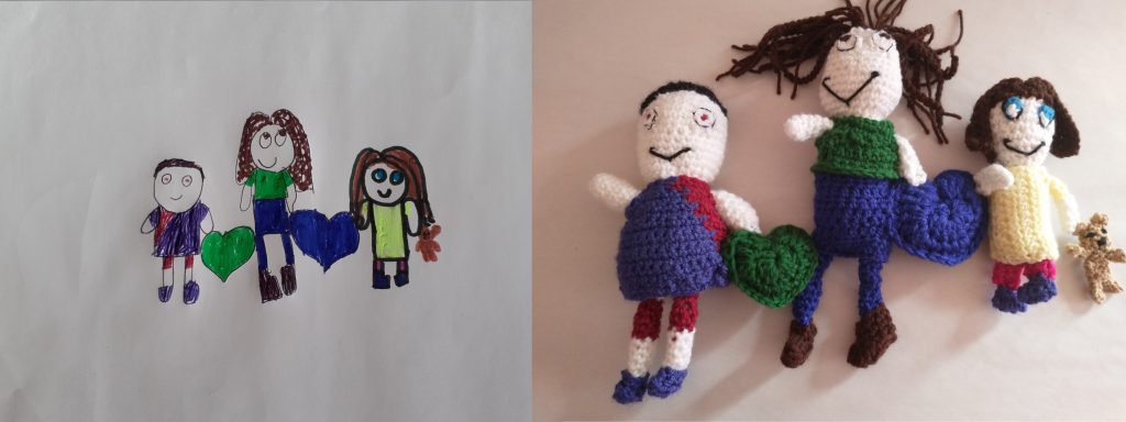 A child's drawing of three children on the left with the crochet toy of the same on the right. There is a little boy on the left wearing a blue and red football shirt. A tall girl in the middle with long brown hair and a green top, blue skirt and leggings and brown boots. The third child is a smaller girl with a yellow dress, pink leggings, blue shoes and brown hair. She is holding a little brown teddy bear. All have big smiles and are joined by a green heart and a blue heart between them.