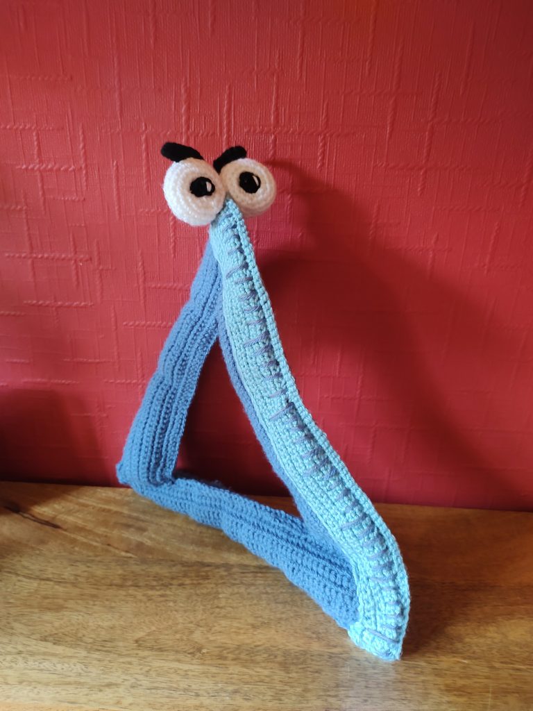 A blue crochet protractor triangle with large white eyes on each side of the top part of the "ruler".He has two thick black eyebrows, one on top of each eye, so it looks like he is frowning.