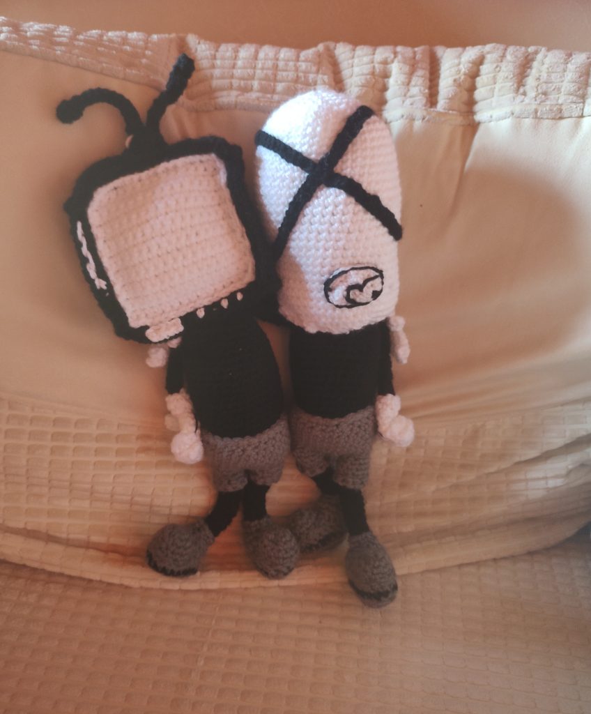 A photo of a cartoon couple with their arms around one another. They both have black tops and grey shorts, and grey cartoon boots and black legs. They have white cartoon gloves on and their fingers are curled on the arms that are hanging down. one has a TV for a head with a black casing and two black antennae and white screen. The other has a white oblong head with two black lines crossing over in the middle. Neither have eyes, but the oblong head one has a black outlined mouth also showing the tongue in outline
