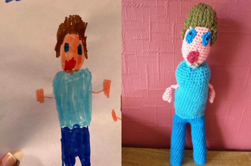 A child's drawing of a man wearing a light blue top, dark blue trousers, with brown hair and a goatee bears. He has blue oval eyes and a pink oval mouth. The crochet toy is to the right of the drawing