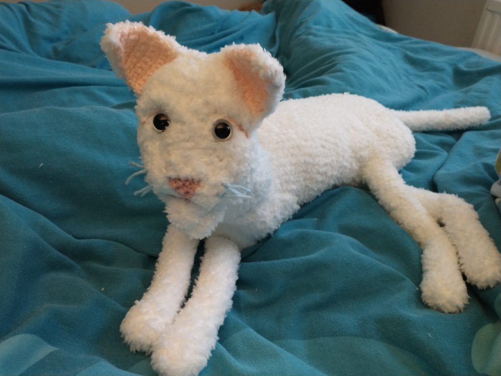 A crochet white cat sitting on a teal bedspread and looking directly at the camera with large grey eyes. He is laying down. His ears and nose are pink.