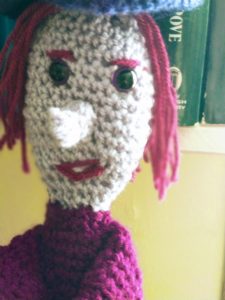 A close-up of the crochet witch's face, showing her purple eyes, long grey nose, straggly purple-pink hair and purple pink eyebrows and lips