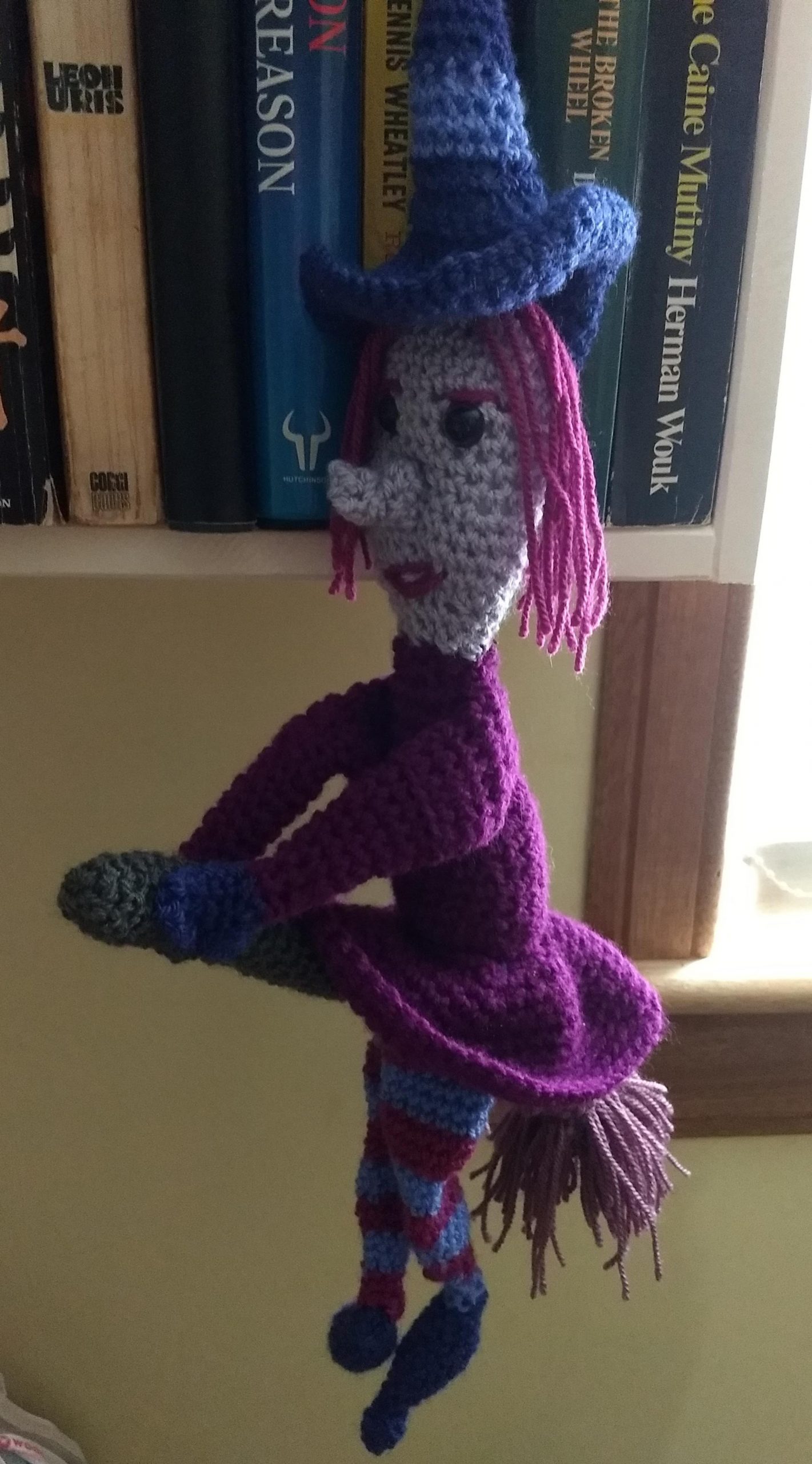 A crochet witch in a deep purple dress and stripey purple witch hat, flying on her brookstick, which is grey with purple-pink bristles, her skin is purple grey and she has a long nose. She is wearing purple striped leggings and long purple shoes.
