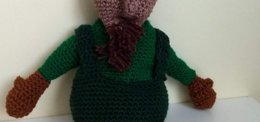 A brown crochet wood sprite wearing a green top and dark green dungarees. He has large brows and green cat eyes that make him look like he is unhappy. He has a dark brown goatee, brown mittens and round brown feet