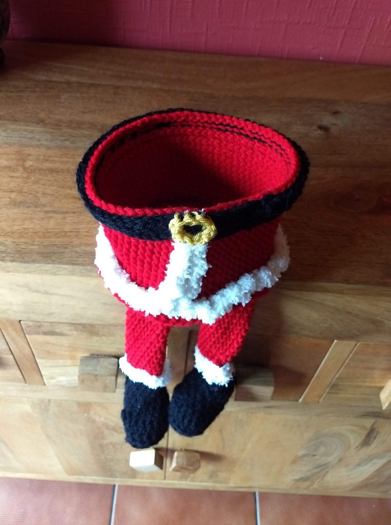 A photo of a little crochet treat basket made to look like the bottom half of Santa sitting on a wooden chest of drawers. The basket is empty and it has two little legs that are hanging over with black boots and fluffy cream edges and red leggings. The basket itself has a black belt with a gold buckle around the top and fluffy trimmings down the middle and around the base