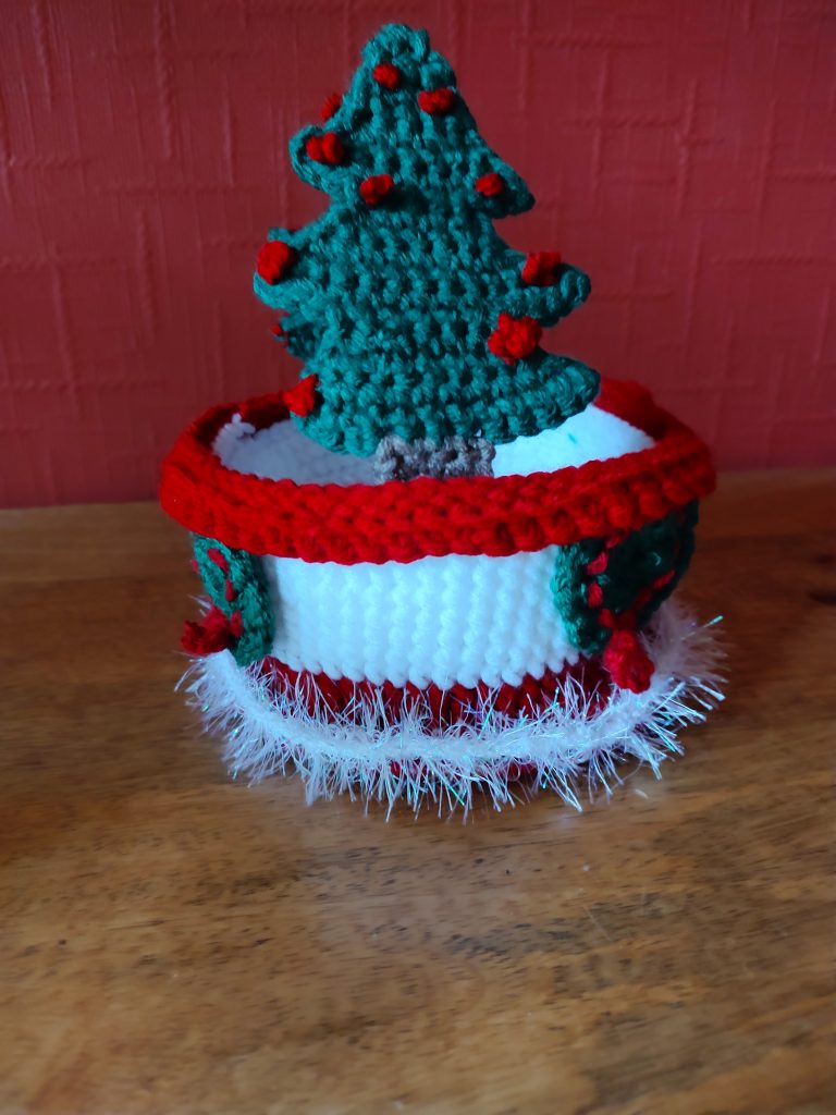 A photo of a crochet Xmas themed basket with a red rim, white body and red bottom around which white tinsel yarn is threaded. On the white body there are green wreaths with red ribbon in them made from crochet chains. The basket has two Christmas trees with red baubles stitched on as the handles. This is the side view showing one of the tree handles straight on