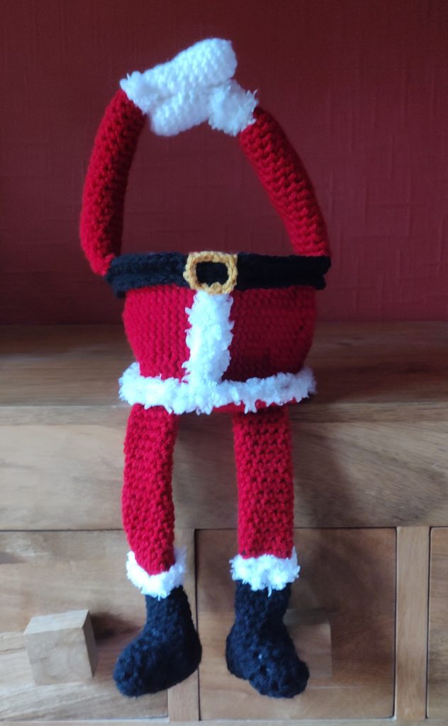 Photo showing the little Santa treat basket empty sitting on the wooden sideboard with his arms above his head for the handle and his legs dangling off the edge,