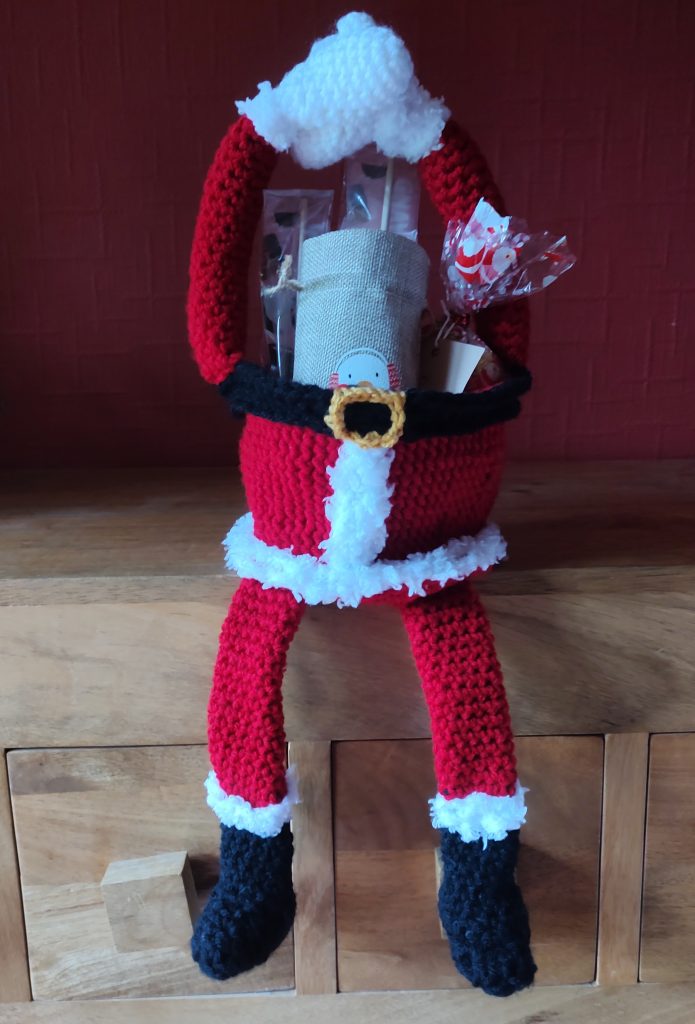 A photo of a little crochet treat basket sitting on a wooden sideboard. The basket has been made to look like Santa's body in red, with fluffy white trimming down the middle and around the base. It also has legs which are dangling over the sides with black boots and white fluffy trimming, and arms with white gloves raised above the body for the handles. It is filled with Christmas goodies