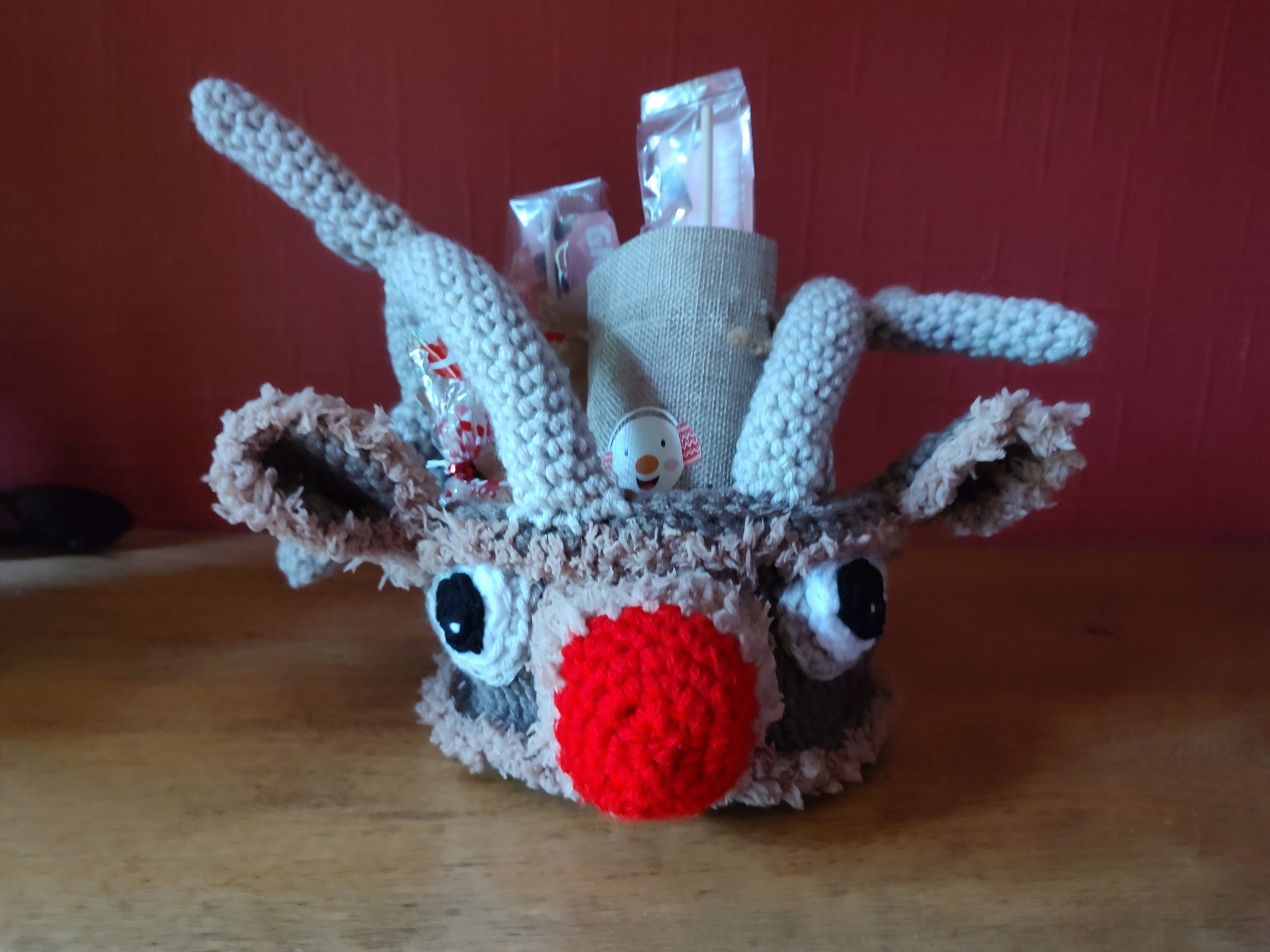 A crochet treat basket made to look like Rudolph the Red Nose Reindeer. The basket is face on showing his large red nose, big eyes and ears. The basket handles are his antlers and the ears, round the nose and bottom and top trim of the basket is made using furry brown yarn. The basket is filled with little Christmas treats