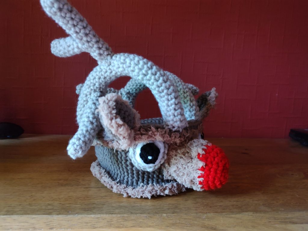 A crochet treat basket made to look like Rudolph the Red Nose Reindeer. The basket is face on showing his large red nose, big eyes and ears. The basket handles are his antlers and the ears, round the nose and bottom and top trim of the basket is made using furry brown yarn. The basket is shown from the side view in this photo.