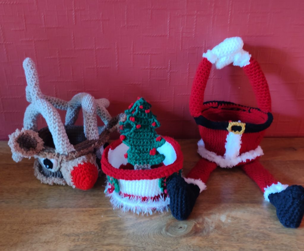 A photo of three crochet treat baskets sitting next to one another on a wooden surface against a red wall. On the right is a Santa version with a Santa suit as the body and two splayed legs with black boots and red trousers with fluffy white trimmings at the ankles. Over the basket the two red arms make the handle, ending in fluffy white trimming at the wrists and white mittens. In the middle is a smaller basket with a white middle around which green Christmas wreaths with red ribbons made from crochet chains are displayed. It has two green Christmas trees with red baubles as the handles and a white tinsel like trimming round the base. ON the left it a basket made to look like Rudolph the Red Nose Reindeer with a red nose, fluffy trimming, two big eyes and fluffy trimmed ears and antlers that form the handles of the basket.