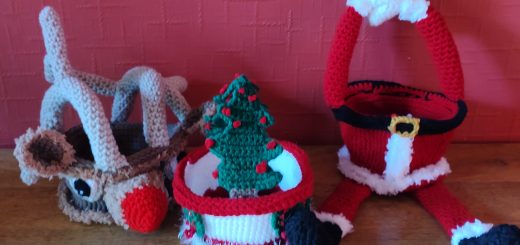 A photo of three crochet treat baskets sitting next to one another on a wooden surface against a red wall. On the right is a Santa version with a Santa suit as the body and two splayed legs with black boots and red trousers with fluffy white trimmings at the ankles. Over the basket the two red arms make the handle, ending in fluffy white trimming at the wrists and white mittens. In the middle is a smaller basket with a white middle around which green Christmas wreaths with red ribbons made from crochet chains are displayed. It has two green Christmas trees with red baubles as the handles and a white tinsel like trimming round the base. ON the left it a basket made to look like Rudolph the Red Nose Reindeer with a red nose, fluffy trimming, two big eyes and fluffy trimmed ears and antlers that form the handles of the basket.