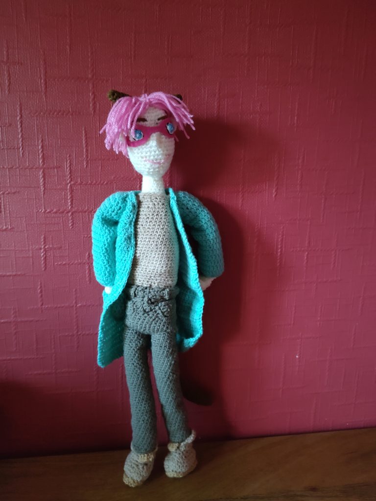 A crochet doll standing on a wooden surface next to a red wall. The doll has pink floppy hair, blue eyes and pink glasses and is wearing a tan shirt, green jacket and khaki trousers. He also has light brown boots and a dark brown tail and pointy ears. He has his hands in the pockets of his jacket and is looking to the right.