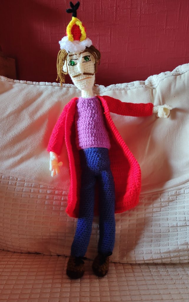A crochet doll of a king standing on a cream chair in front of a red wall. The King is wearing a fur trimmed red and gold grown with a black cross on top. He has green eyes, long brown hair and a brown pencil moustache. He is wearing a purple top, a long red coat, blue trousers and brown lace up boots. He is pointing to the right and his other arm is down at his side.