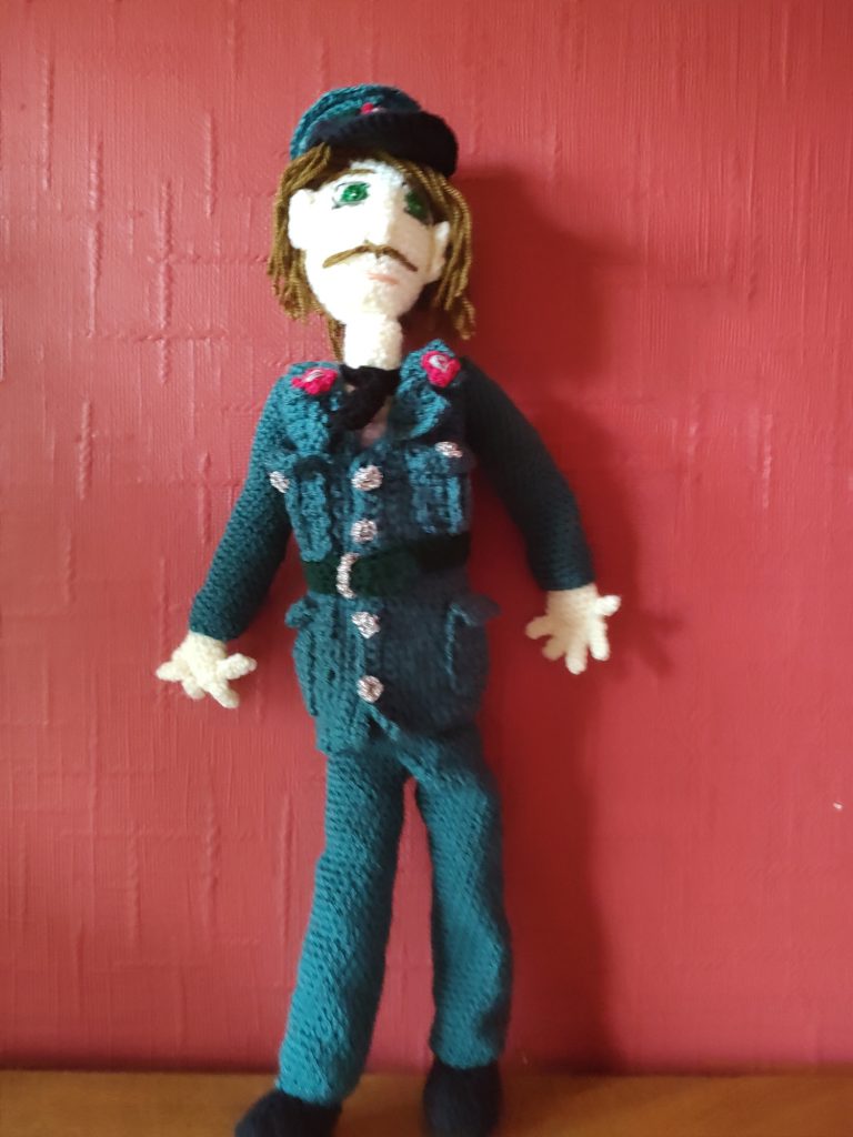 A crochet doll standing on a wooden surface next to a red wall wearing an Ulster police uniform which is a green-blue colour with silver buttons, a darker green belt and buttoned top pockets and fold over hip pockets. The trousers are the same colour, as is the hat, which has a black brim. He is wearing black shoes and a black tie. The lapels and front of the cap bear red flashes with the logo of a harp on them. His hair is long and brown and he has a brown pencil moustache and green eyes.