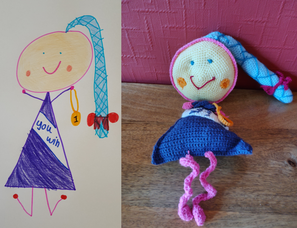 A child's drawing of a little girl with the matching crochet toy to the left. The little girl is wearing a triangular dark blue dress with a white sash and the words "You Win" on it in blue. She has a large round yellow head with a pink outline and a large light blue pony tail wound around with darker blue "string". The pon y tail has a red bow at the end of it. She is holding a golden medallion with the number 1 on it and she wears a big happy pink smile above her two orange round cheek spots. She has blue stick arms and pink stick legs.