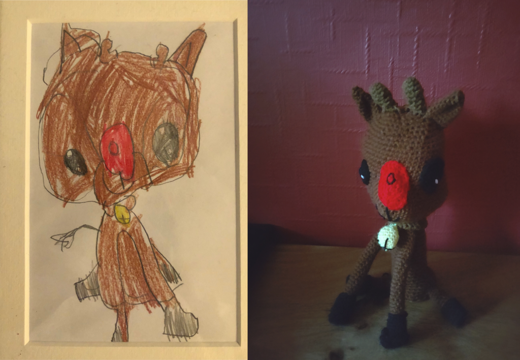 A child's drawing of a reindeer, and to the right the crochet toy made from it. The reindeer in the drawing is shown from the front. He has a large brown head, brown ears, a red nose and large black eyes and a black snout outlined in black with a slight smile. His body is smaller and also brown with black boot like feet. He has a skinny black, feathered tail and is wearing a golden yellow bell around his neck.