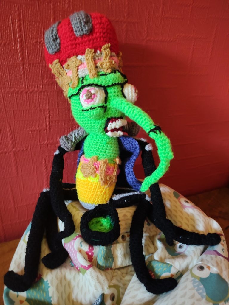 A crochet toy of a bin weevil. It has a green head with a Frankenstein monster like cap with grey staples in it and bandages. It has a very long green, pointed nose and large bulging eyes and teeth. It has skinny black legs and arms and is holding a little black pot with green bubbling goo in it. The body is stripey yellow and orange with more bandages on it and it is wearing a blue cardigan.
