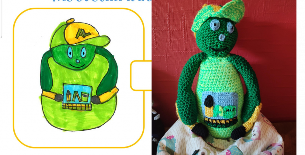 A child's drawing of a round boy, wearing a lime green top with long, dark green sleeves and a baseball cap with a yellow front with the letter A on it and a green peak. He has light blue eyes and nose and a large black smile. He has a picture, representing school tools such as a protractor, on his belly. The crochet toy version of the boy is next to this, sitting on a little owl beanbag next to a red wall.