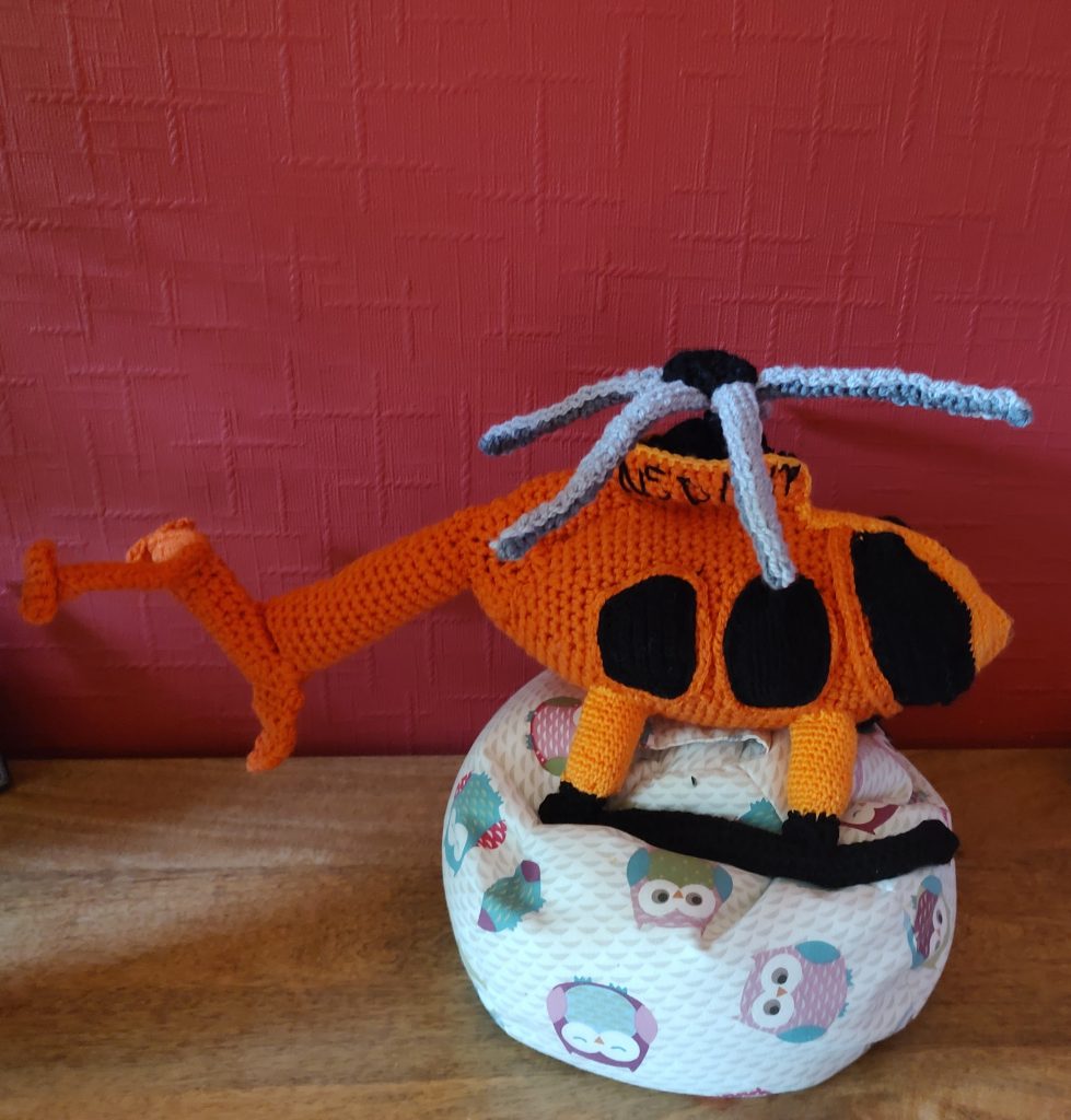 An orange crochet helicopter sitting on a little owl beanbag next to a red wall. The helicopter has grey rotor blades and three black windows visible from this side view. It has fat lighter orange legs on black runners and a large tail with various fins sticking out of it. There is black writing on the top of the fuselage just under the rotor blades.