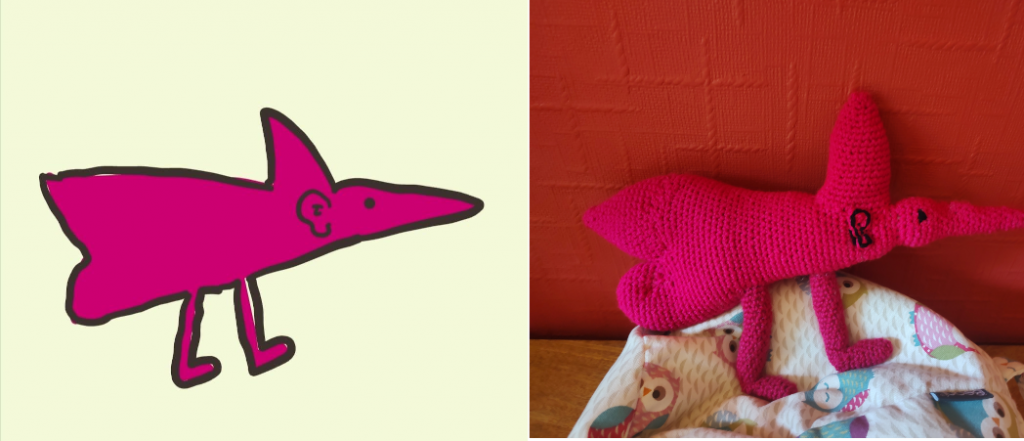 A drawing of a raspberry coloured creature in side view that looks a little like a triangle mouse with a shark fin on its back. It has two legs and a black dot eye, small black ear and a long pointed snout. The crochet toy version of the drawing is on the right, sitting on a little own beanbag next to a red wall.