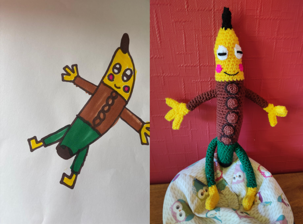 A child's drawing of a banana man, wearing a brown, long-sleeved top with brown buttons down the front and green trousers. It has a large yellow face with white eyes and slitted black pupils. It is smiling and has two pink cheeks. It has a black stalk on its head and yellow three fingered hands, and yellow feet. The crochet version of the drawing is on the right, sitting on a little owl cushion next to a red wall.