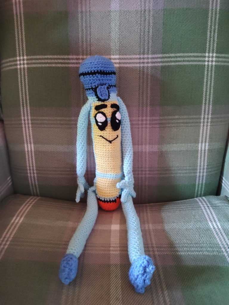 A photo of an epi pen crochet toy sitting on a green tartan chair. It has light blue arms and legs with darker blue boots and a pen top. It has a yellow body in front and sports large eyes and a little smile