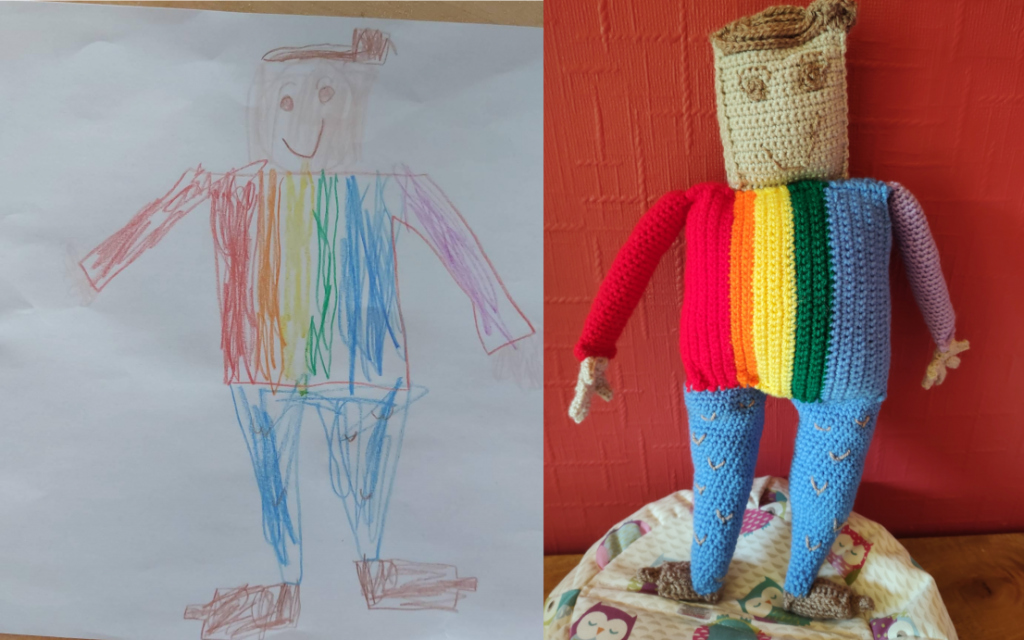 A child's drawing of a square man with a rainbow top which runs from blue on the right, through green, yellow, orange and red on the left. |The right arm is purple and the left red. He also has a square tan head with a swirl of brown hair ending in a cowlick on the right. He has brown eyes and eyebrows and a brown smile. He is wearing blue trousers with little brown v shapes all over them and has brown shoes. The crochet toy version of the drawing is on the right sitting on a little owl beanbag next to a red wall.