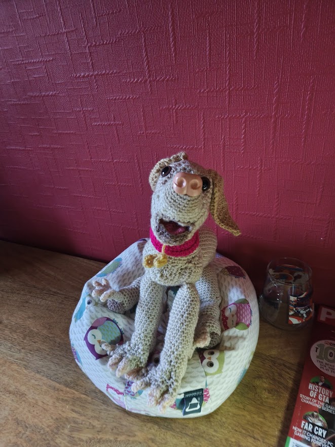 A crochet tan dog sitting on a little owl cushion next to a red wall. The dog has a large pale pink nose, brown eyes and long darker tan ears. It's mouth is open in a puppy smile and it has its head cocked slightly to one side. It's big front paws are visible with skin-coloured nails on each of the four toes. The dog has a pink collar with a gold bone on it.