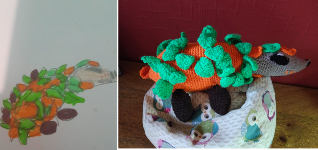 A child's drawing of "Leafhog" an orange hedgehog like creature which is covered in green leaves. It has small flat oval brown feet and a tan snout with three orange markings on each side. There are two black nostrils on the snout and it has large oval black eyes with white highlights in them. The view of the drawing is showing Leafhog from the side with its snout facing right. On the right is the crochet toy of the drawing in the same position. It is sitting on a little owl beanbag next to a red wall.