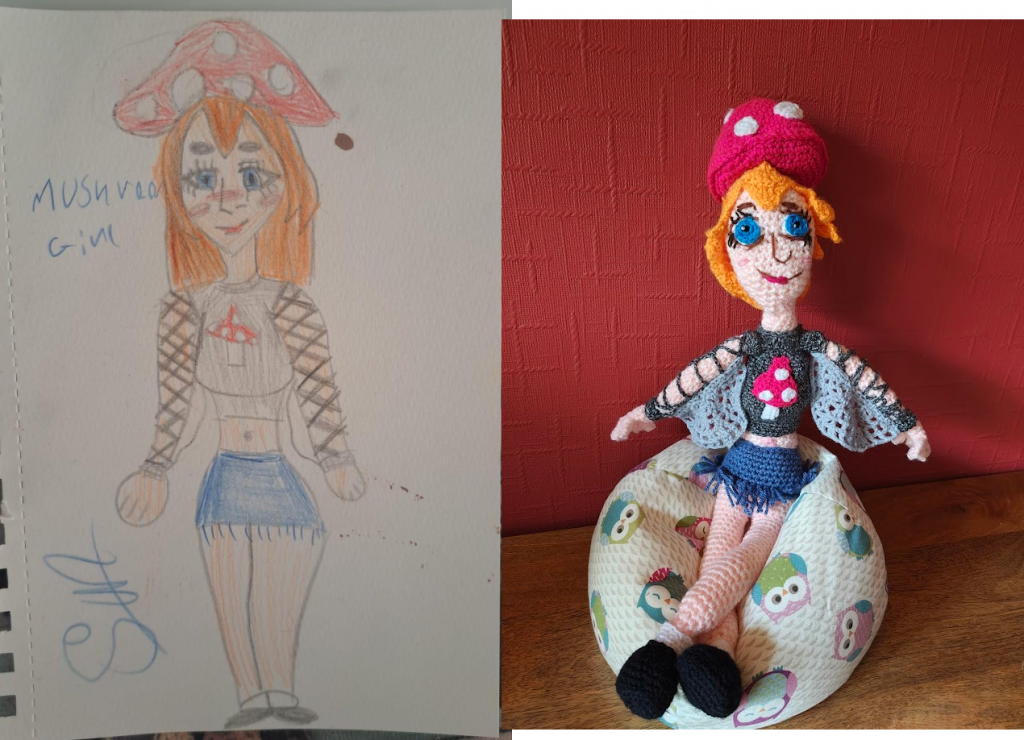 A child's drawing of a girl with long orange hair wearing a pink hat with white spots in the shape of a mushroom. She has large blue eyes with long lashes and is smiling with her mouth closed. She is wearing a dark grey top that has strings zigzagging down the arms leaving her pink arms bare underneath. The top has wings underneath the arms a bit like spider webs. There is another pink mushroom with white spots and a white stem on her top. She is wearing a denim mini skirt with tasselled edges. Her pink legs are bare and she is wearing black shoes with short white socks. The crochet version of the drawing is on the right, sitting on a little own beanbag next to a red wall with her arms outstretched to show the "wings" and her legs crossed over at the ankles.