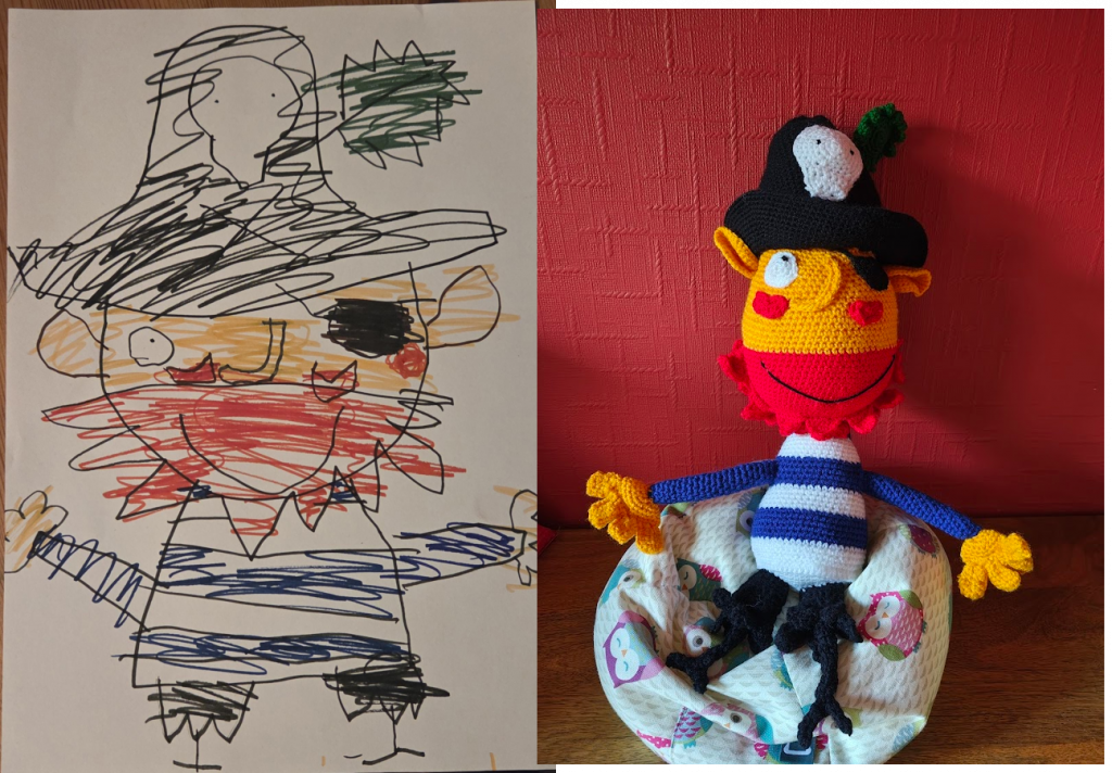 A child's drawing of a pirate wearing a large black hat with a white skull on the front and a leaf sticking out from it. He has a large yellow orange head and a wavy red beard and red cheeks. He is wearing a large black smile and an eyepatch over one eye, while the other is white with a small black pupil. He has a blue and white striped long sleeved top on and yellow orange hands with four fingers. He has two very skinny peg legs sticking out from black ragged shorts. On the right is the crochet toy version of the drawing sitting on a little owl beanbag next to a red wall.