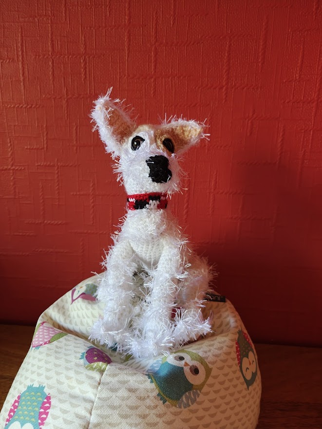 A crochet white terrier dog with wiry fur sitting on a little owl beanbag next to a red wall. The dog has brown eyes and a black nose and mouth. It has triangular ears which are perked up as it looks at the camera. It is wearing a tartan collar