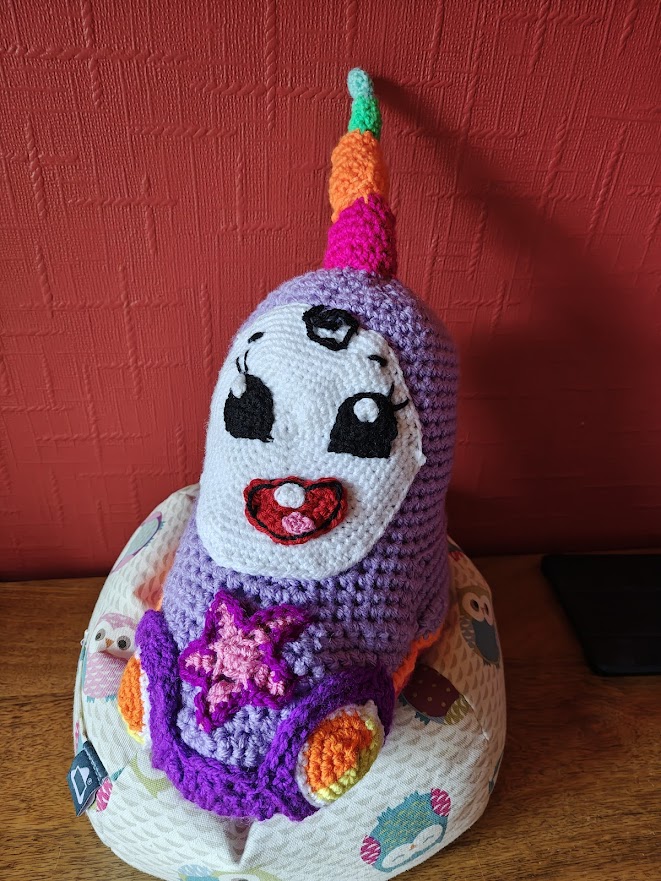 A crochet toy of the baby uni car from a children's cartoon. It has a large round white face on its dome shaped lavender head, with a wide open red mouth showing a little pink tongue and one white baby tooth on the top. It has large black eyes with white irises and long black eyelashes, and a black curl on its forehead. On the bonnet is a pink and purple star and the front headlights, which are orange and yellow are visible as is the dark purple bumper. It has a large horn like protrusion on its head that spirals from pink to orange to green to light blue-green.
