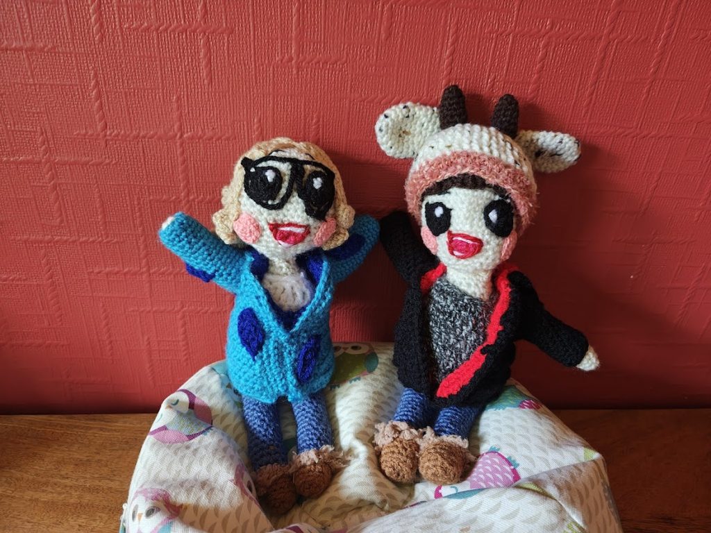 Two little crochet dolls, both smiling widely, sitting side by side on a little owl beanbag next to a red wall. The one on the left has her arms thrown up in the air. She has blonde hair and is wearing black glasses, a blue hooded coat with dark blue leaf motives on it, blue jeans and brown booties with fluffy ankle outlines. She is wearing a white top underneath her jacket. On the right the other doll has her arms spread out. She is wearing a tweedy beanie hat with a pink tweed trim, floppy ears and dark brown horns in the middle. She is wearing a black hooded jacket with red trim, jeans and boots the same as the first doll and a mottled grey top.